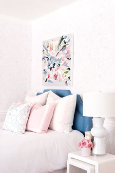 a white bed topped with lots of pillows next to a lamp and painting on the wall