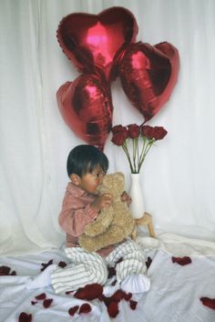 a little boy holding a teddy bear in front of some heart shaped balloons and flowers