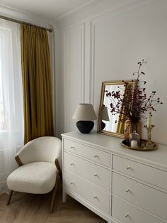 a white dresser topped with a mirror next to a lamp and a vase filled with flowers