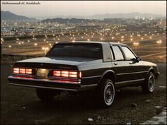 a black car parked on top of a dirt field next to a cityscape