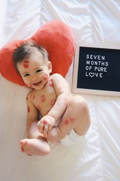 a baby laying on top of a white bed next to a red heart shaped pillow