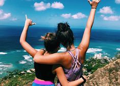 two women standing on top of a hill with their arms in the air and looking at the ocean