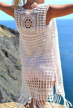 a woman wearing a white crochet dress standing on top of a cliff overlooking the ocean