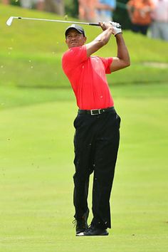 a man in red shirt and black pants hitting a golf ball with his club on the green