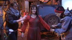 a woman in a red dress is holding out her hand to another person who is standing next to a jeep