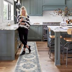 a woman standing in the middle of a kitchen with blue cabinets and an area rug