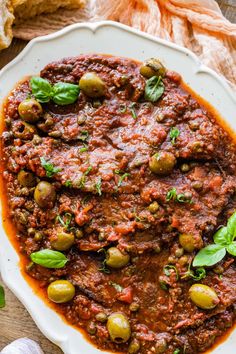 a white plate topped with meat and olives covered in marinara sauce next to garlic bread