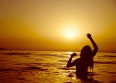 a woman standing in the ocean at sunset raising her arms up to the sun above her head