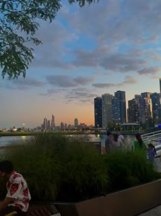 people are sitting on benches near the water in front of tall buildings and skyscrapers