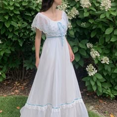 a woman wearing a white dress standing in front of some bushes and flowers with her eyes closed