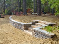 a stone wall in the middle of a dirt field with trees and benches behind it