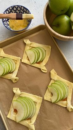 four pieces of apple pie sitting on top of a cookie sheet next to bowls of apples