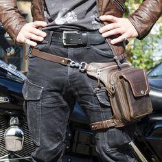 a man wearing a leather jacket and belt with his hands in his pockets, standing next to a motorcycle