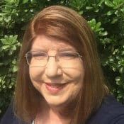 a woman with glasses smiling for the camera in front of green bushes and trees behind her