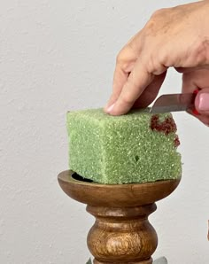 a person cutting into a green cake on top of a wooden stand next to a white wall