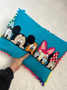 a person is holding a blue pillow with mickey and minnie mouses on it, which has pom - poms around the edges