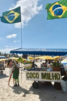 two people standing in front of a food cart on the beach with flags flying above them