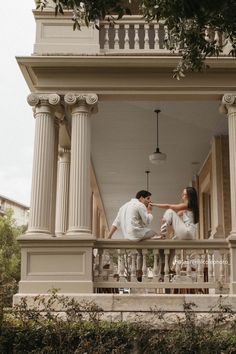 a man and woman sitting on the porch of a house with columns in front of them