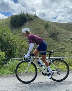 a woman riding a bike down a street next to a lush green hillside covered in clouds