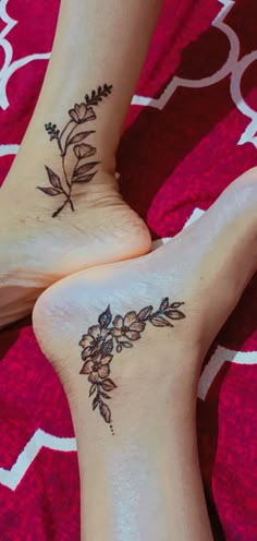 two feet with flower tattoos on them sitting on a red bed spread and one has her foot up to the other side