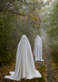 two white ghost statues standing in the middle of a leaf covered path surrounded by trees