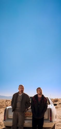 two men standing next to each other in front of a white car on the desert