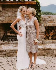 two women standing next to each other in front of a stone fire place with greenery