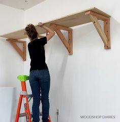 a woman standing on top of a ladder next to a wooden shelf above a white wall