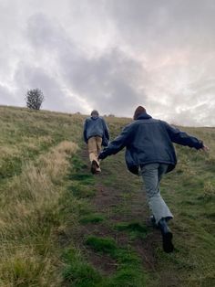 two people walking up a grassy hill on a cloudy day with one holding the other's hand