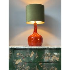 a red vase sitting on top of a green dresser next to a lamp with a green shade