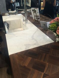 a white sink sitting on top of a wooden counter next to a mirror with people in the background