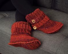 two red knitted boots sitting on top of a gray couch next to a woman's leg