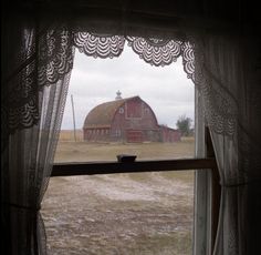 a red barn sitting in the middle of a field next to a window with lace curtains