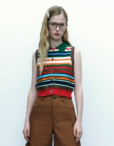 a woman with glasses standing in front of a white wall wearing brown pants and a striped shirt