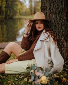 a woman sitting on the ground next to a tree wearing a brown hat and coat