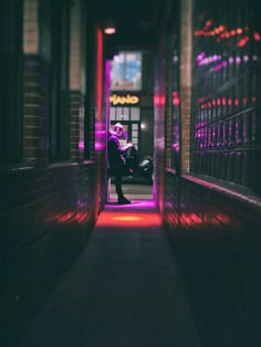 a person sitting on a bench in an alley way with red light coming from the windows