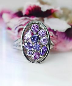 a ring with purple and white stones on it sitting next to some pink flower petals