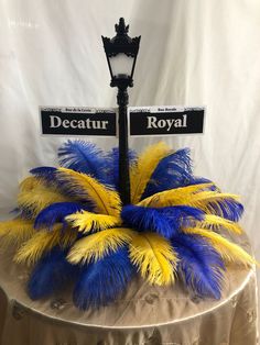 a table topped with a lamp post covered in blue, yellow and white feathers next to a sign that reads decatur royal