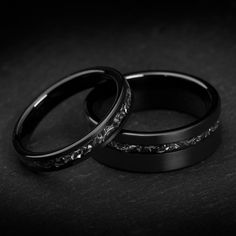 two black wedding rings sitting next to each other on top of a dark surface,