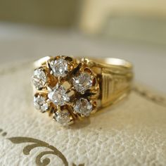 This dazzling Victorian statement ring features a cluster of old mine cut diamonds in vibrant 18 karat yellow gold. Seven (7) .25 carat diamonds are set in a flower cluster on the front of the ring. The diamonds are near colorless and eye clean, bringing a striking contrast and brightness to the warm yellow gold. From the profile, ornate Victorian crown baskets hold the collection of diamonds. The shoulders of this cocktail ring feature unique engravings finished on the tapered 18K yellow gold b Yellow Gold Diamond Cluster Ring, Heirloom Yellow Gold Cluster Wedding Ring, Antique Gold Cluster Diamond Ring, Vintage Yellow Gold Cluster Ring, Gold Cluster Ring With Rose Cut Diamonds For Wedding, Vintage Gold Flower Ring, Extravagant Rings, Victorian Crown, Italian Ring