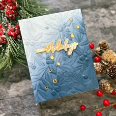 a blue christmas card with gold foil lettering and pine cones on the table next to it