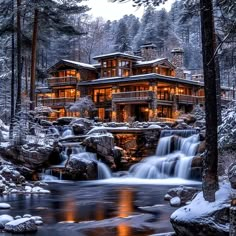 a house in the woods with a waterfall running through it and lights on at night