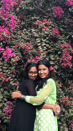 two women hugging each other in front of pink flowers on a wall covered with vines