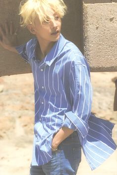 a young man with blonde hair wearing a blue shirt and jeans standing in front of a stone wall