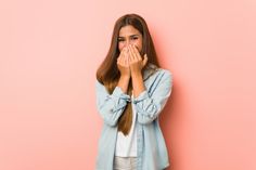 a young woman covering her mouth with both hands while standing against a pink wall looking at the camera