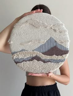 a woman holding up a piece of art made out of yarn and fabric with mountains in the background