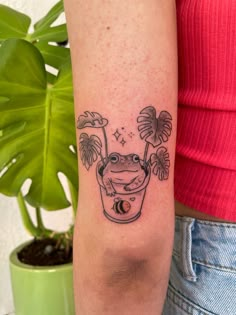 a small frog in a glass tattoo on the right inner arm and leg, next to a potted plant