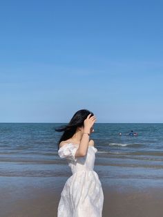 a woman in a white dress standing on the beach looking at the ocean with her eyes closed