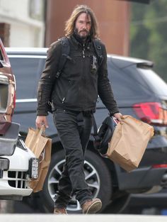 a man with long hair and beard carrying shopping bags walks down the street in front of parked cars