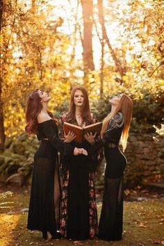 three women in long black dresses are standing together and looking at an open book while the sun shines through the trees behind them